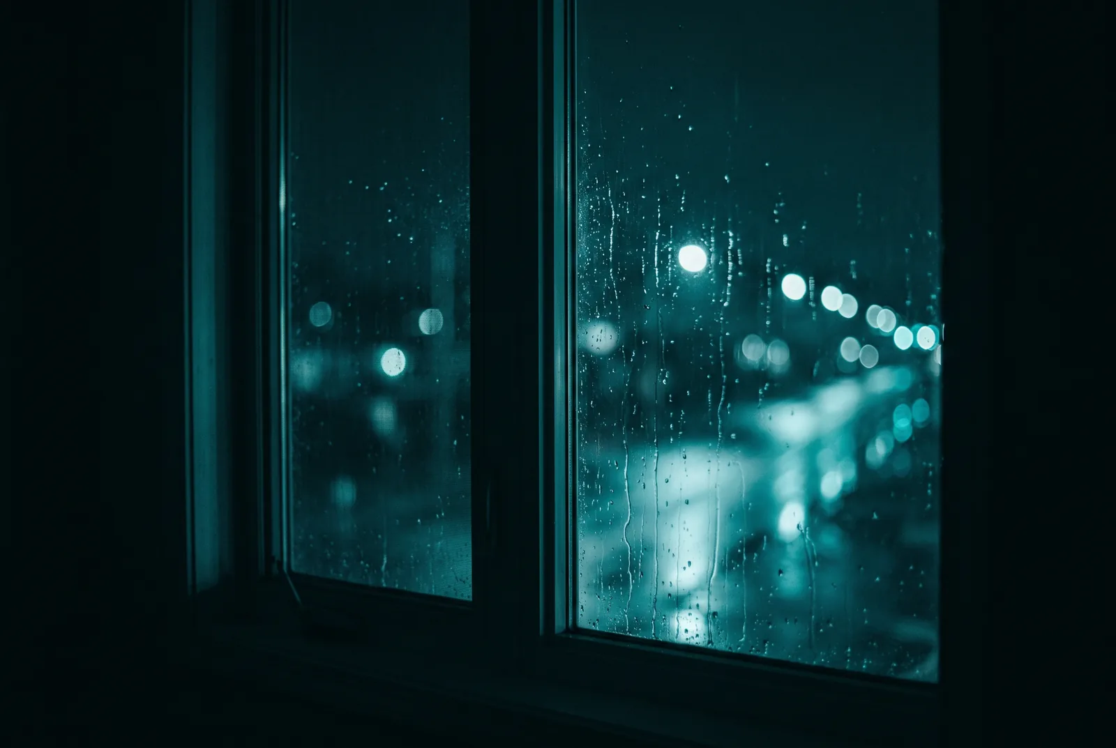 A tall window at night from inside a dark room, rain running down the glass, teal street lights out of focus beyond